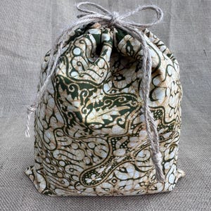 May include: A drawstring pouch made of fabric featuring a dark green and beige batik pattern with intricate swirling designs. The bag is cinched at the top with a thick, natural-coloured jute twine cord tied into a bow, creating a gathered closure.