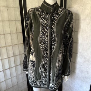 May include: A long-sleeved, button-down jacket with a high collar. The jacket features a mix of olive green, black, and white patterns, including zebra stripes and abstract designs. The jacket has a tailored fit and a dark trim.