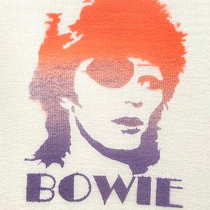 Puede incluir: Una camiseta blanca con una silueta morada y naranja de la cara de David Bowie y la palabra "BOWIE" impresa en morado.