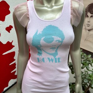 May include: A pink tank top with a blue graphic of David Bowie's face and the word "BOWIE" printed on the front. The tank top has pink tulle ruffles on the shoulders.