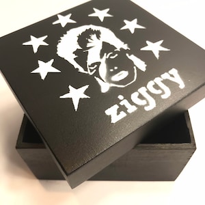 May include: A black wooden box with a white silhouette of David Bowie as Ziggy Stardust and the text "Ziggy" on the lid. The lid is open and the box is surrounded by a white background.
