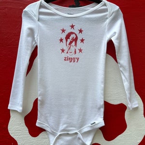 May include: White long-sleeve baby onesie with a red print of David Bowie's face with the text "ziggy" underneath. The print is surrounded by a circle of red stars.