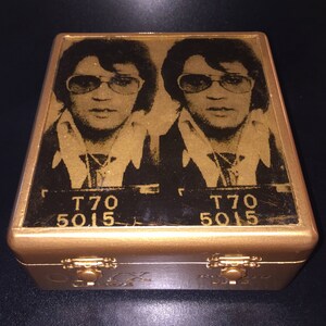 Op de afbeelding: Een gouden houten doos met een scharnierend deksel. Het deksel heeft een zwart-witfoto van Elvis Presley met de tekst "T70 5015" aan beide kanten.