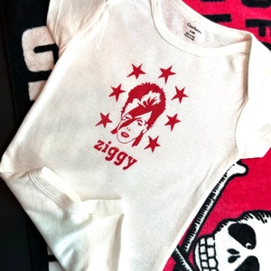 Może przedstawiać: Kremowe body niemowlęce z czerwoną grafiką twarzy otoczonej gwiazdami i napisem "ziggy". Ubranko ma krótkie rękawy i jest wykonane z miękkiego materiału. Widoczna jest metka marki Gerber.
