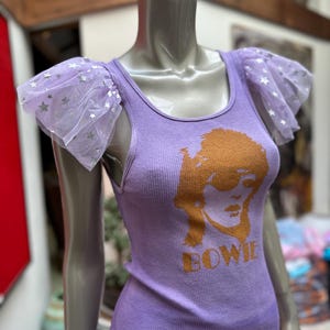 May include: Purple tank top with a graphic print of David Bowie's face and the word "BOWIE" underneath. The tank top has sheer purple tulle sleeves with silver stars.