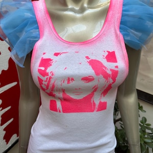 Op de afbeelding: Een wit tanktop met een roze afbeelding van een vrouwen gezicht. Het tanktop heeft blauwe tule ruches op de schouders.
