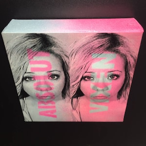 May include: A close-up of a painting featuring two women with their faces partially obscured by pink and blue paint. The women have long, dark hair and are looking directly at the viewer.