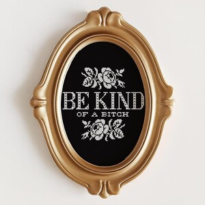 Op de afbeelding: Een ovaal, goudkleurig ingelijst kruissteekwerk met de woorden "BE KIND OF A BITCH" in het wit, met bloemmotieven boven en onder de tekst. De achtergrond is zwart.