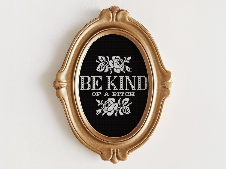 Op de afbeelding: Een ovaal, goudkleurig ingelijst kruissteekwerk met de woorden "BE KIND OF A BITCH" in het wit, met bloemmotieven boven en onder de tekst. De achtergrond is zwart.