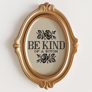 Op de afbeelding: Een ovale, goudkleurige lijst met kruissteek. De tekst "BE KIND OF A BITCH" in zwart is omgeven door zwarte bloemmotieven. De lijst heeft een sierlijk, decoratief ontwerp.