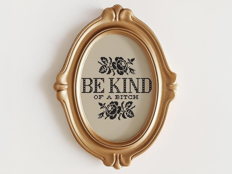 Op de afbeelding: Een ovale, goudkleurige lijst met kruissteek. De tekst "BE KIND OF A BITCH" in zwart is omgeven door zwarte bloemmotieven. De lijst heeft een sierlijk, decoratief ontwerp.