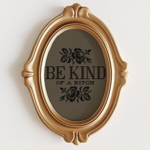 Op de afbeelding: Goudkleurige, ovaalvormige fotolijst met de tekst "BE KIND OF A BITCH" in zwart. De lijst heeft een sierlijk ontwerp en de tekst is omgeven door bloemmotieven. De achtergrond is donkergrijs.