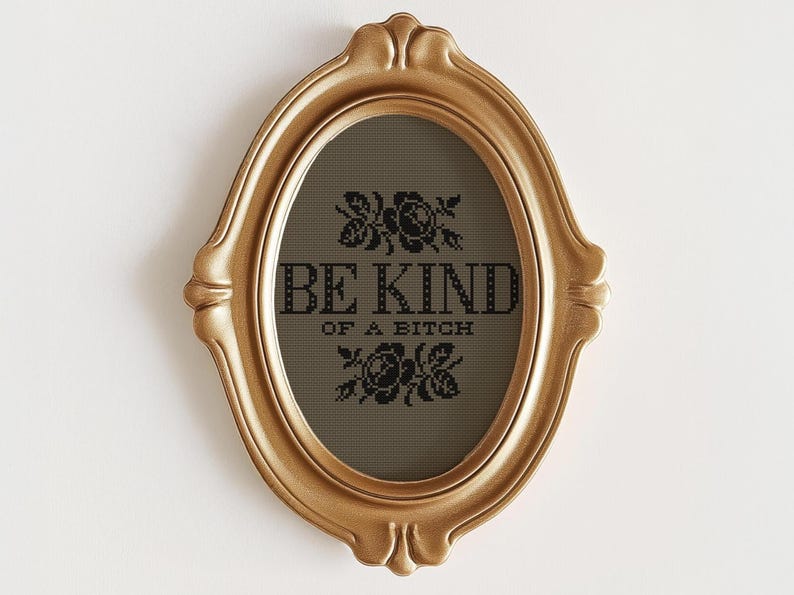 Op de afbeelding: Goudkleurige, ovaalvormige fotolijst met de tekst "BE KIND OF A BITCH" in zwart. De lijst heeft een sierlijk ontwerp en de tekst is omgeven door bloemmotieven. De achtergrond is donkergrijs.