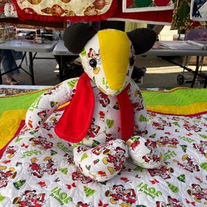 Pode incluir: Um brinquedo de pel&uacute;cia branco e vermelho com tema do Mickey Mouse com uma cabe&ccedil;a amarela e preta. O brinquedo est&aacute; usando uma echarpe vermelha e est&aacute; sentado em um edredom branco com um padr&atilde;o do Mickey Mouse. O edredom tem as palavras "Season of Joy!" impressas.