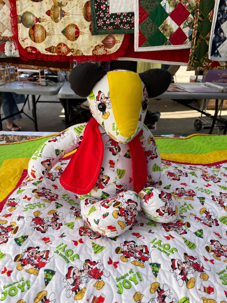 Pode incluir: Um brinquedo de pel&uacute;cia branco e vermelho com tema do Mickey Mouse com uma cabe&ccedil;a amarela e preta. O brinquedo est&aacute; usando uma echarpe vermelha e est&aacute; sentado em um edredom branco com um padr&atilde;o do Mickey Mouse. O edredom tem as palavras "Season of Joy!" impressas.