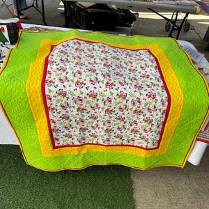 Pode incluir: Um edredom branco com uma estampa de Natal vermelha e verde. O edredom tem uma borda amarela e verde. O edredom est&aacute; dobrado e sentado em uma superf&iacute;cie verde.