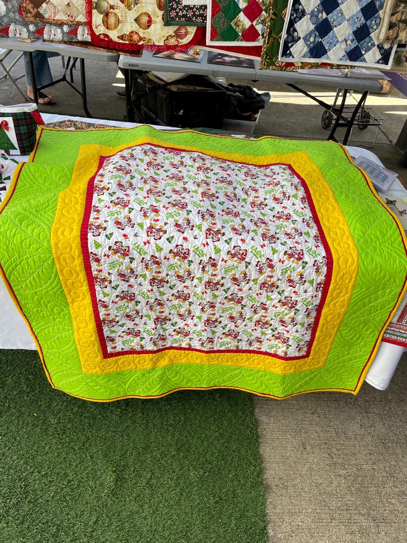 Pode incluir: Um edredom branco com uma estampa de Natal vermelha e verde. O edredom tem uma borda amarela e verde. O edredom est&aacute; dobrado e sentado em uma superf&iacute;cie verde.