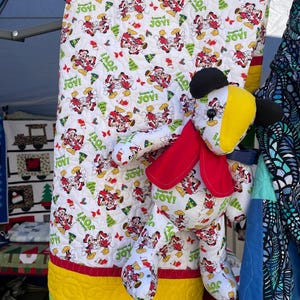 Pode incluir: Uma colcha branca com uma borda vermelha, verde e amarela. A colcha apresenta um padr&atilde;o repetitivo de Mickey Mouse e Minnie Mouse com roupas de Natal. Um brinquedo de pel&uacute;cia do Mickey Mouse est&aacute; na frente da colcha.