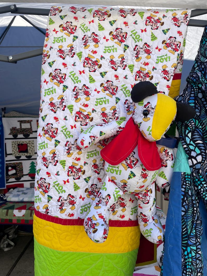 Pode incluir: Uma colcha branca com uma borda vermelha, verde e amarela. A colcha apresenta um padr&atilde;o repetitivo de Mickey Mouse e Minnie Mouse com roupas de Natal. Um brinquedo de pel&uacute;cia do Mickey Mouse est&aacute; na frente da colcha.