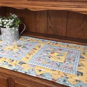 May include: A yellow and blue quilted table runner with a farmhouse theme. The runner features a patchwork design with various patterns and images of gardening tools, birds, and houses. The runner is displayed on a wooden table with a small galvanized watering can filled with white flowers.