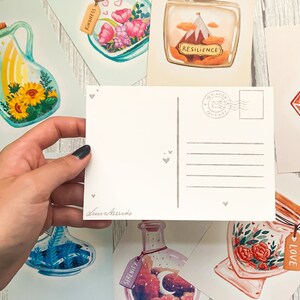 May include: A hand holds a blank postcard with a gray heart design, surrounded by watercolor art prints. The prints feature jars with various themes, including sunflowers, flowers, and a mountain scene. Each jar has a tag with a word.