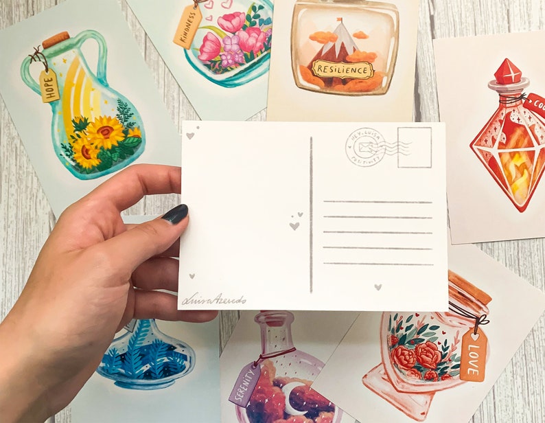 May include: A hand holds a blank postcard with a gray heart design, surrounded by watercolor art prints. The prints feature jars with various themes, including sunflowers, flowers, and a mountain scene. Each jar has a tag with a word.