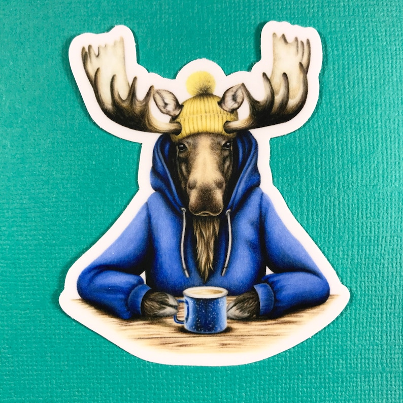 May include: A moose wearing a blue hoodie and a yellow beanie holds a blue mug with a white rim. The moose is sitting at a wooden table.