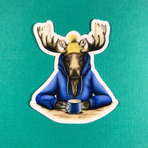 May include: A moose wearing a blue hoodie and a yellow knit hat is holding a blue mug. The moose is sitting at a table with a white surface.