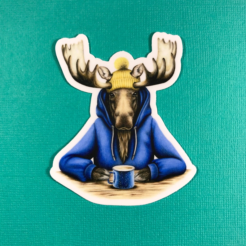 May include: A moose wearing a blue hoodie and a yellow knit hat is holding a blue mug. The moose is sitting at a table with a white surface.