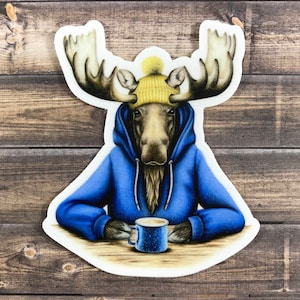 May include: A moose wearing a blue hoodie and a yellow beanie holds a blue mug with a brown beverage. The moose is sitting at a wooden table.