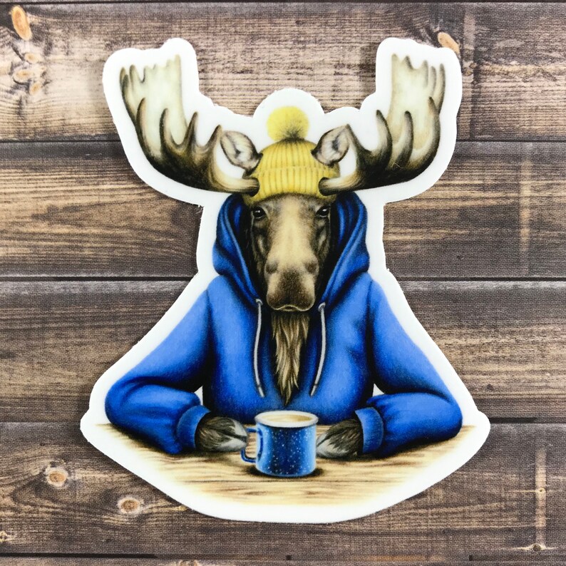 May include: A moose wearing a blue hoodie and a yellow beanie holds a blue mug with a brown beverage. The moose is sitting at a wooden table.