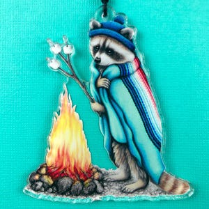 May include: A raccoon wearing a blue hat and a colorful blanket sits by a campfire with marshmallows on a stick. The raccoon is looking at the marshmallows.