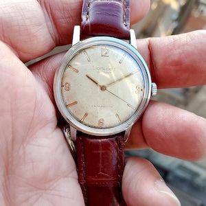 May include: A silver wristwatch with a white face and black hands. The watch has a brown leather band and says "Cortebert Sport" and "Antimagnetic" on the face.