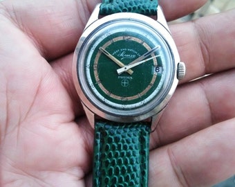 May include: A green-faced wristwatch with a silver case and a green leather band. The watch face has a white dial with black markings and the words "West End Watch Co. Prima" and "Saxon" printed on it.