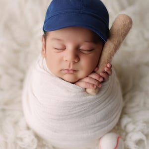 May include: A newborn baby wrapped in a white blanket, wearing a navy blue baseball cap. A small felt baseball bat rests near the baby's face, and a felt baseball with a red seam is next to the baby.