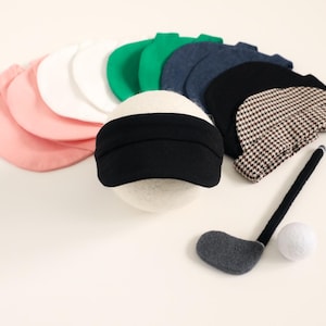 May include: A collection of miniature golf-themed items, including a golf club, a golf ball, and several small hats in various colors such as pink, white, green, blue, black, and a houndstooth pattern. The hats are arranged in a semi-circle.