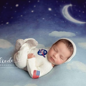 Puede incluir: Un bebé recién nacido vistiendo un traje de astronauta blanco con un parche de la bandera estadounidense en la manga, durmiendo sobre un fondo azul y blanco con forma de nube con una luna creciente en la esquina superior derecha.
