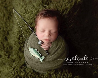 May include: A newborn infant wrapped in a dark olive green swaddle blanket, holding a miniature black fishing rod with a small green and white fish lure attached to the line. The text rosalinda PHOTOGRAPHY WWW.ROSALINDAPHOTOGRAPHY.COM is visible.
