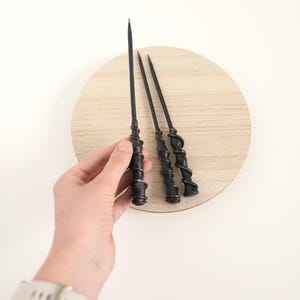 May include: Three black wands with spiral detailing are arranged on a light wood-grain circular surface. One wand is held by a hand. The wands have pointed tips and are likely for cosplay or themed events.