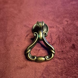 Könnte beinhalten: Ein antiker Schubladengriff aus Messing mit einem verzierten Design. Der Griff hat eine kreisförmige Grundplatte und einen geschwungenen Griff mit aufwendigen Details. Die Hardware befindet sich vor einem weinroten Hintergrund.