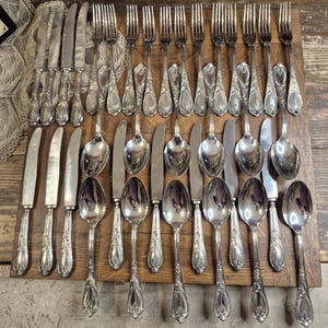 May include: A collection of vintage silverware, including knives, forks, and spoons, arranged on a wooden surface. The silverware has ornate, floral-like detailing on the handles. The spoons, forks, and knives are made of a shiny, silver-colored metal.
