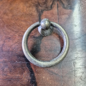 Peut inclure: Gros plan d'une poignée de tiroir en bronze antique avec une poignée circulaire et un petit détail en forme de boule. La quincaillerie est montée sur une surface en bois foncé, mettant en valeur la patine vieillie du métal.