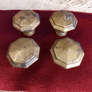 May include: Four antique brass cabinet knobs with an octagonal design. The knobs have a weathered, aged appearance, with visible patina and slight discoloration. They are arranged on a deep red velvet surface.