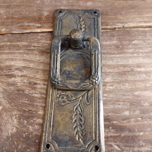May include: Antique brass door pull with a floral design and a ring handle. The pull is mounted on a rectangular plate.