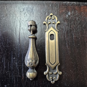 Könnte beinhalten: Zwei antike Schrankgriffe in Messing-Optik. Ein Griff ist eine lange, rechteckige Platte mit verzierten Details. Der andere Griff ist ein tropfenförmiger Griff mit einem dekorativen Ring.
