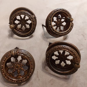 Pode incluir: Quatro botões de armário de latão antigo com um design decorativo, circular e vazado. Cada botão tem uma puxador de anel e um parafuso decorativo.