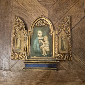 Pode incluir: Um tríptico com um painel central apresentando uma pintura de uma mulher segurando uma criança, ladeado por dois painéis laterais com relevos de figuras. A moldura é dourada com topo arqueado, volutas detalhadas e uma base azul.