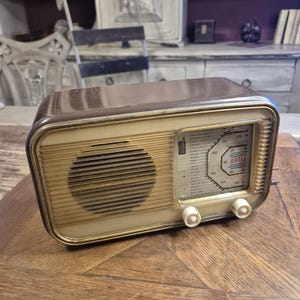 Op de afbeelding: Een vintage tafelradio met een donkerbruine kunststof behuizing en een crèmekleurig voorpaneel. De voorzijde is voorzien van een cirkelvormig luidsprekerrooster aan de linkerkant en een rechthoekige afstemschaal met de merknaam ARTIA in het rood.