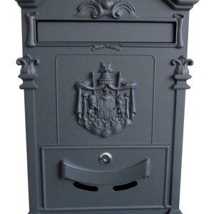 May include: A dark grey metal mailbox with a decorative design and a slot for mail. The mailbox has a keyhole and a handle for opening the door.