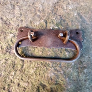 May include: A rusty metal drawer pull with a decorative, ornate design. The pull has a rectangular shape with rounded edges and a slightly curved handle.